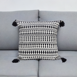 Square throw pillow with black and white geometric pattern and black tassels on each corner, placed centered on a light gray couch with a plain white background.