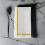 White linen napkin with mustard yellow embroidered border and white fringed edges, folded on a gray marble surface with dark veining.