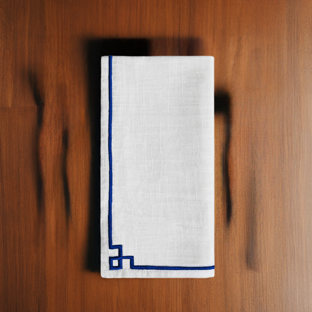 White fabric napkin with a deep blue embroidered border and corner detail, displayed folded on a warm brown wooden surface.