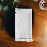 White linen napkin with fringe edges and simple black embroidered border, folded on a wooden table with leafy greenery and a glass nearby.