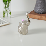 Small silver-finished elephant ring holder with trunk raised, placed on white surface near a plant in a glass vase and wooden base with textured dark object.