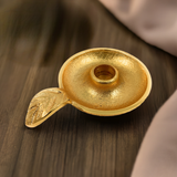 Round gold-toned taper candlestick holder dish with textured finish and a leaf-shaped handle, placed on a dark wooden surface.