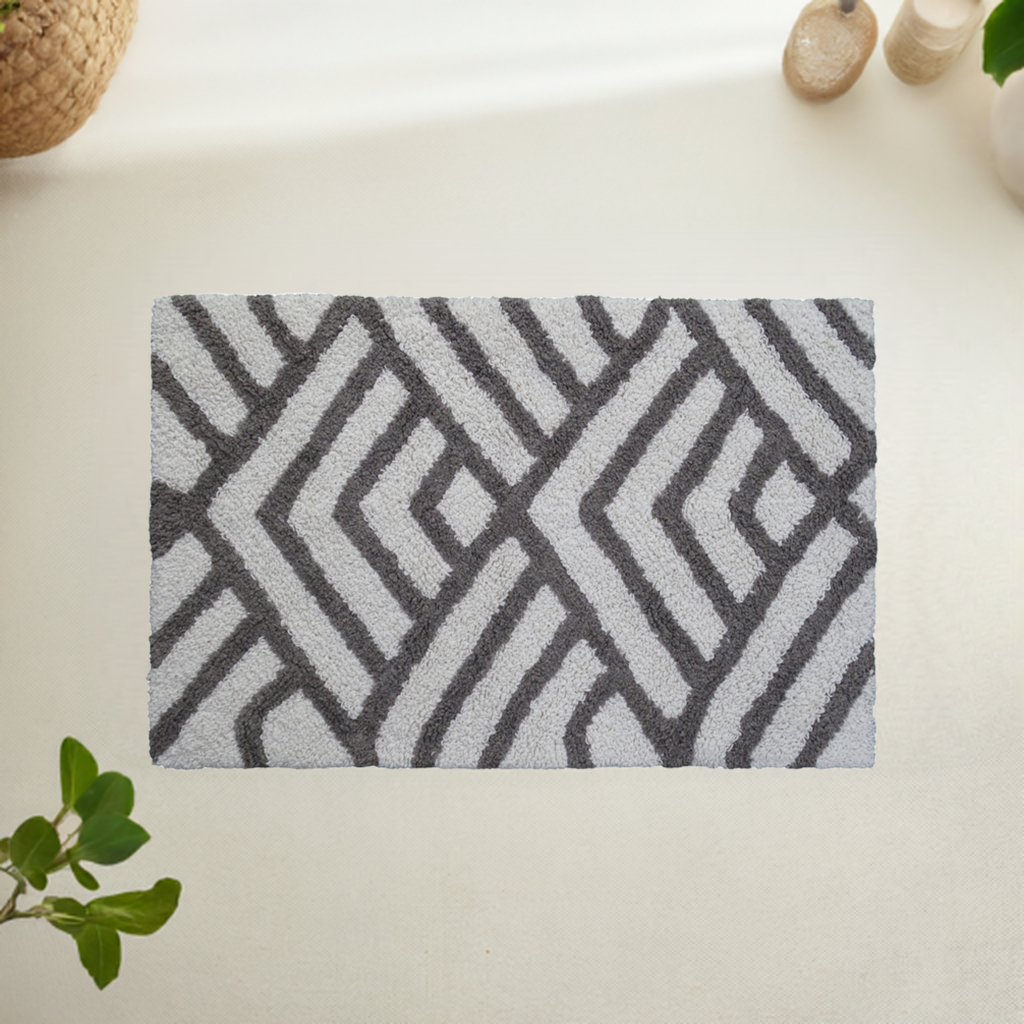 Rectangular bath rug with grey and ivory geometric diamond pattern laid on a light floor, framed by green plant leaves and natural textures.