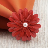Red floral napkin ring with layered petal design and a central pearl-like bead, holding an orange cloth napkin on light wood surface.