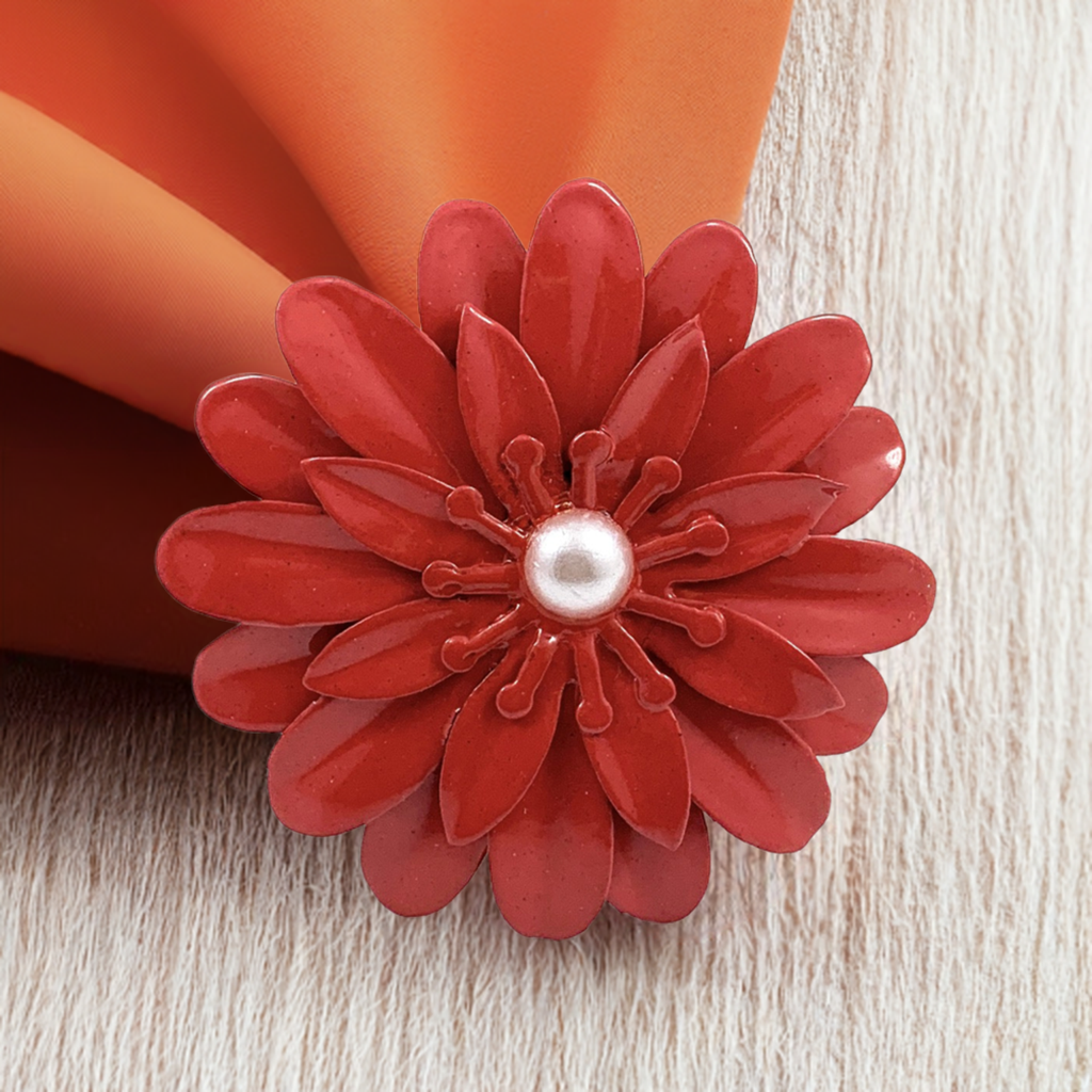 Red floral napkin ring with layered petal design and a central pearl-like bead, holding an orange cloth napkin on light wood surface.
