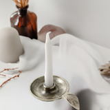 Silver-toned metal candlestick holder with a small leaf accent and a white taper candle, set on a white draped fabric with a brown glass bottle and candles in the background.