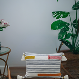 Rectangular white couch pillow with horizontal gray stripes, colorful braid embroidery in mustard yellow, pink, and orange, and tassels on each corner, resting on a wooden surface beside green plants.