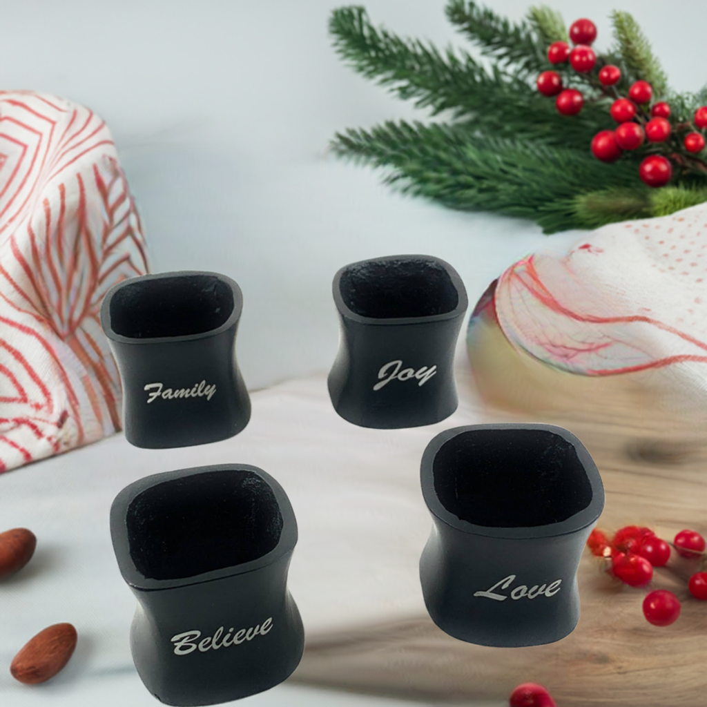 Set of four black napkin rings with white script reading Family, Joy, Believe, and Love, arranged with pine sprigs and red berries on a neutral surface.