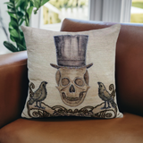 Beige throw pillow embroidered with a skull wearing a tall, dark top hat and flanked by two black birds with curled tails, resting on a brown leather chair.