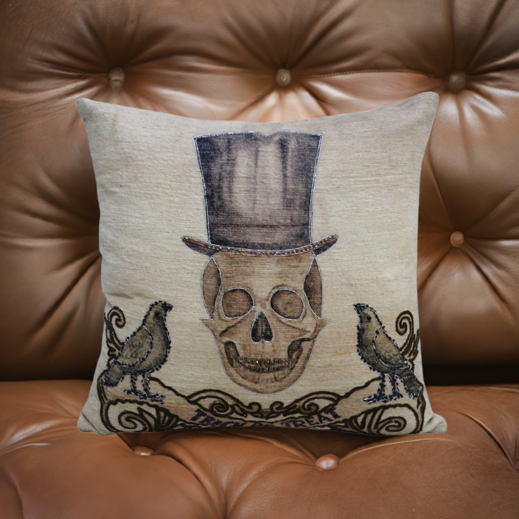 Beige throw pillow with embroidered skull wearing a tall top hat, flanked by two black birds perched on scroll-like designs, placed on a brown tufted leather surface.