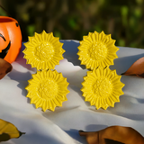 Set of four bright yellow sunflower-shaped napkin rings with textured centers, arranged on a white cloth with soft natural light and autumn leaves nearby.