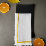 White linen napkin with yellow embroidered border and fringed edges, shown flat on a gray surface with two orange slices nearby.