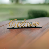 Golden cursive word "Believe" mounted on a slim black base, shown on a wooden surface with a blurred green garden background.