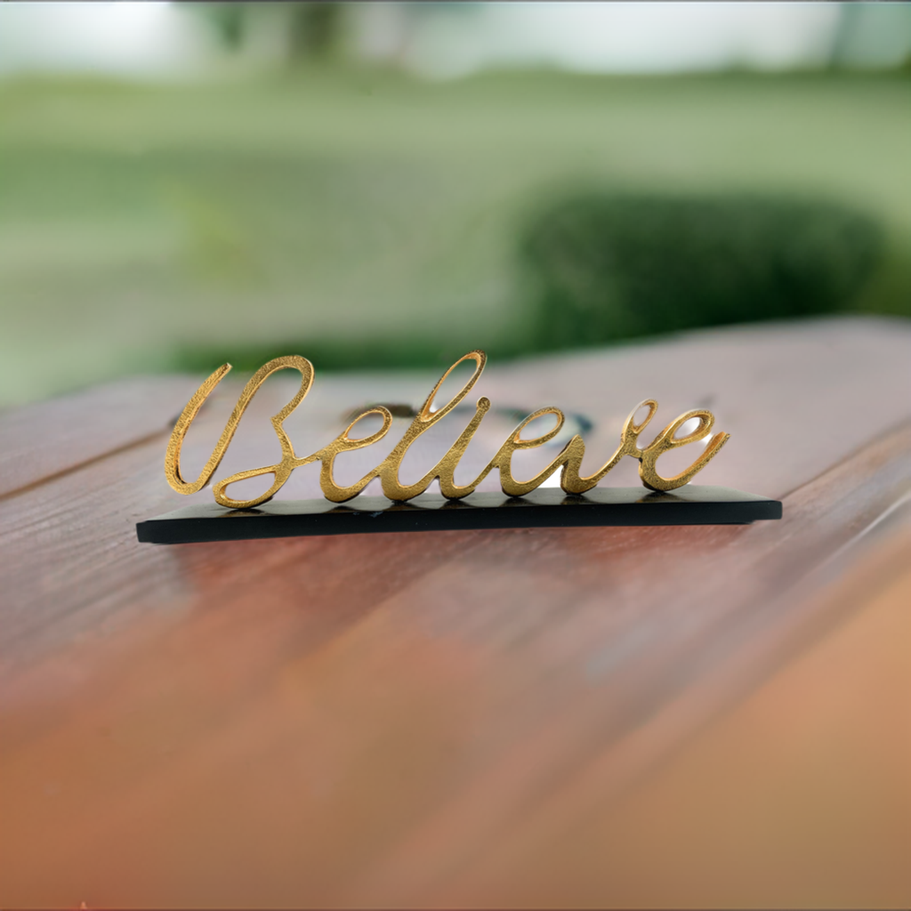 Golden cursive word "Believe" mounted on a slim black base, shown on a wooden surface with a blurred green garden background.