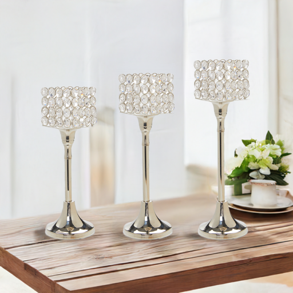 Set of three silver aluminum candle holders with cylindrical tops adorned by clear round crystals, arranged by height on a wooden table beside a small white flower pot and plate.