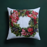 Square white throw pillow with a painted Christmas wreath featuring red poinsettias, pine cones, and green foliage at its center on a dark green background.
