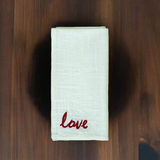 Cream-colored cloth napkin with red cursive 'love' embroidery folded neatly on a dark wooden surface with natural grain patterns.