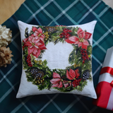Square pillow with a Christmas wreath design featuring red poinsettias, pinecones, and green foliage on a white background, placed on a dark green plaid fabric.