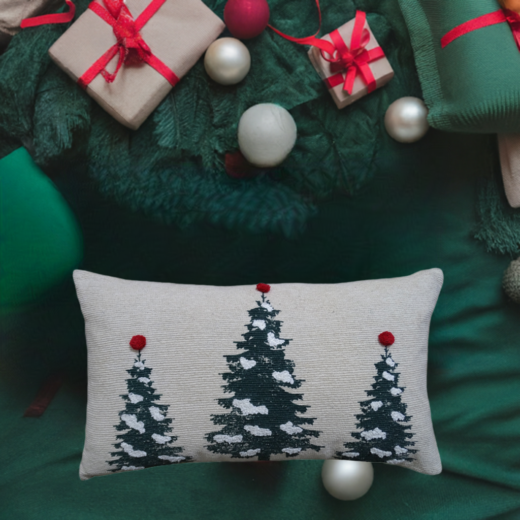Rectangular cream-colored throw pillow featuring three dark green Christmas trees with white snow accents and red ornaments on top, placed on green fabric with gifts and ornaments nearby