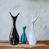 Three ceramic deer figures in glossy black, teal, and white with elongated necks and minimalist design, placed on a wooden surface against a textured light wall.