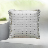 Square white decorative pillow with small black horizontal dash pattern and fringed edges on a light gray fabric chair with soft background.