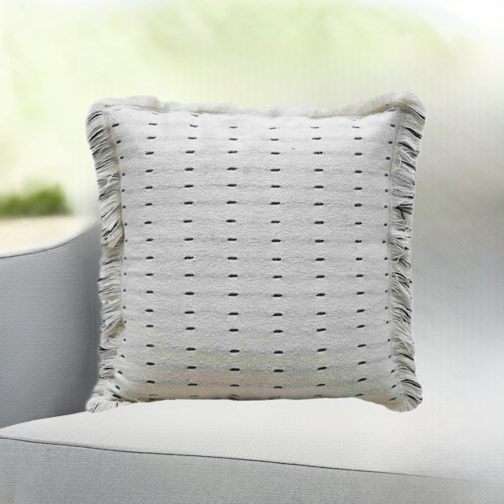Square white decorative pillow with small black horizontal dash pattern and fringed edges on a light gray fabric chair with soft background.