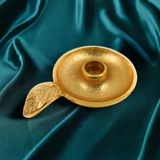 Small gold candlestick holder dish with textured surface and attached leaf-shaped handle, placed on teal satin fabric.