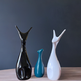 Three glossy ceramic deer figures with elongated necks and minimalist shapes in black, teal, and white on a light surface against a dark background.