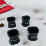 Set of four matte black ceramic napkin rings with curved square shapes, each engraved with white script words: Family, Joy, Believe, and Love, displayed on a white marble surface.