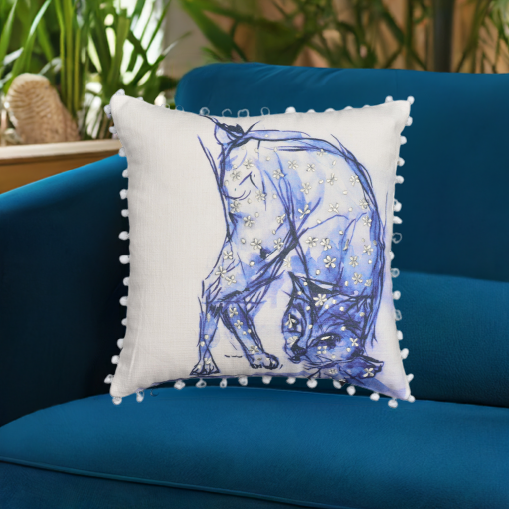 Blue throw pillow with embroidered cat motif, pom-poms and tassels on a white surface