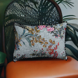 Rectangular decorative pillow with floral and butterfly patterns in pink, red, yellow, and green hues on a pale blue-gray background, edged with black pom-poms.