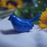 Glossy blue bird figurine with carved feather details, positioned on a soft white surface with blurred green foliage and yellow flowers in background.