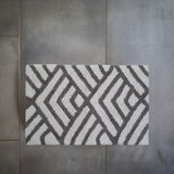 Rectangular bath rug with a geometric pattern of dark grey lines forming diamond shapes on an ivory background, placed on large grey floor tiles.