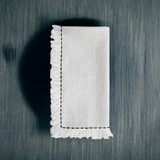 White linen napkin with black embroidered border and fringed edges, neatly folded on a dark textured surface