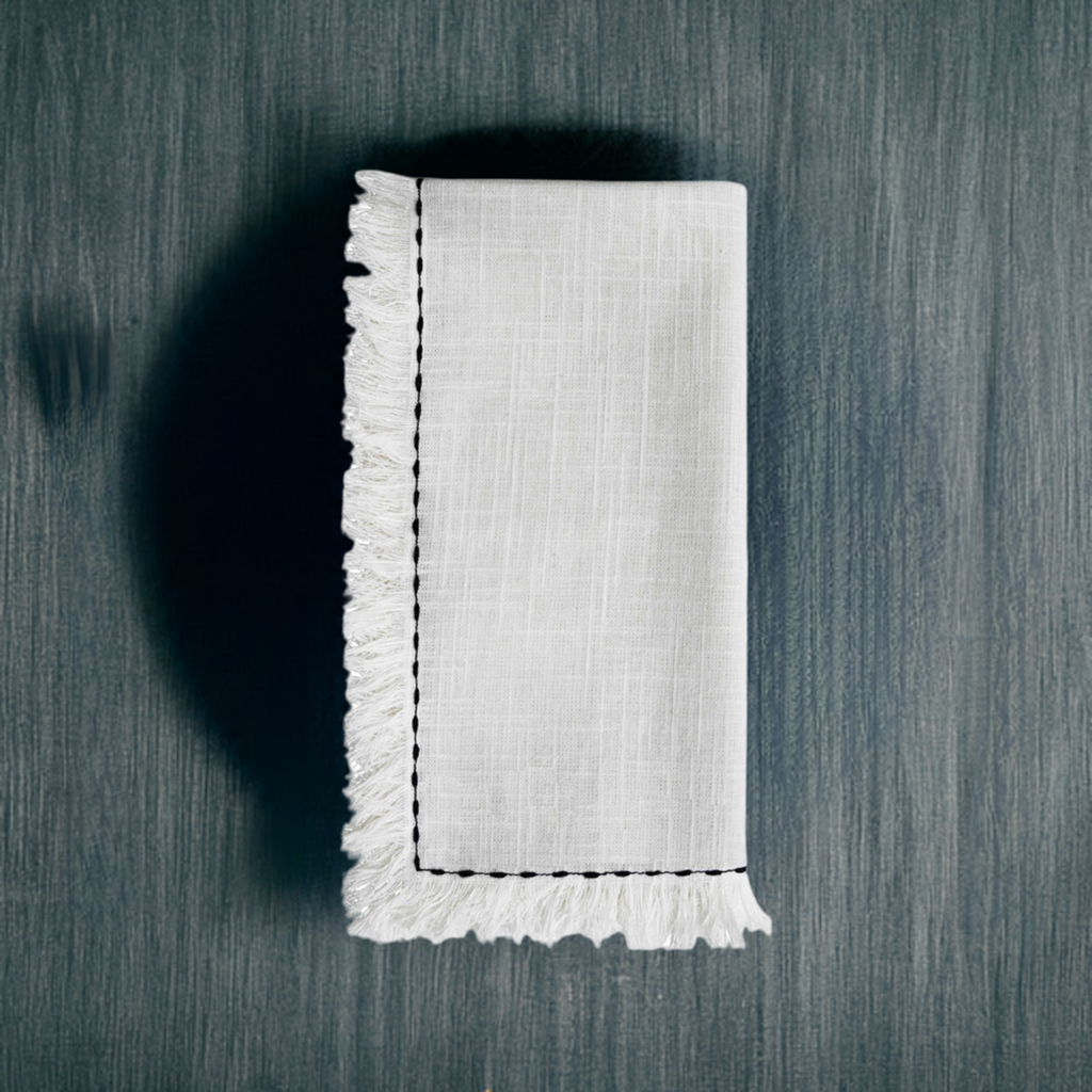 White linen napkin with black embroidered border and fringed edges, neatly folded on a dark textured surface