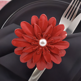Red floral napkin ring with layered petals and a central pearl detail, securing a black napkin placed on a black plate with silver fork and knife