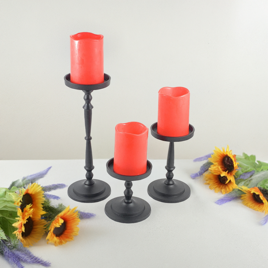 Set of three black metal pillar candle holders of varying heights, each holding a red pillar candle, surrounded by yellow sunflowers and purple flowers on a white surface.