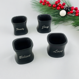 Set of four black resin Christmas napkin rings, each engraved with white words: Family, Joy, Believe, and Love, arranged on a white surface with pine and red berry decoration background.