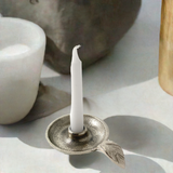 Silver taper candlestick holder dish with textured surface and leaf-shaped handle holding a white candle casting a soft shadow on a light surface.