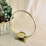 Golden circular metal candle holder on a rectangular base, designed to hold one candle, set against a light fabric backdrop with a white wicker basket and green foliage nearby.