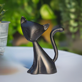 Small black matte cat figurine with elongated neck and curved tail, sitting on a white surface with blurred green plants in the background.