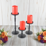 Three black metal pillar candle holders of varying heights with red pillar candles, arranged on a wood floor with floral arrangements on either side.