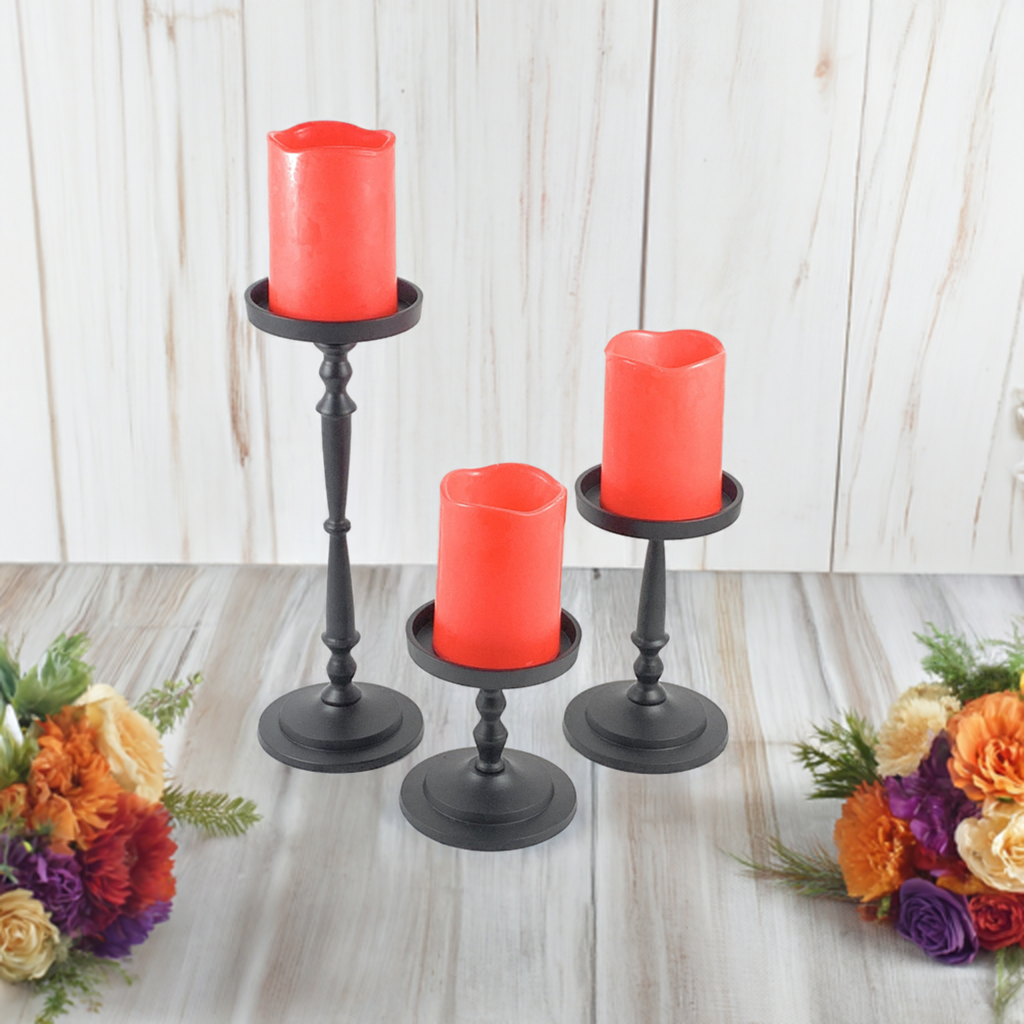 Three black metal pillar candle holders of varying heights with red pillar candles, arranged on a wood floor with floral arrangements on either side.