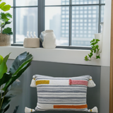 Rectangular couch pillow with gray and white woven stripes accented by horizontal yellow, pink, and rust embroidery braids and tassels on each corner, placed on a chair by a modern window.