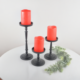 Set of three black metal pillar candle holders in varying heights, each holding a red pillar candle, arranged on a white round table with a small green foliage accent.
