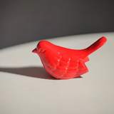 Small red bird figurine with textured feather details casting a shadow on a light gray surface with dark background.
