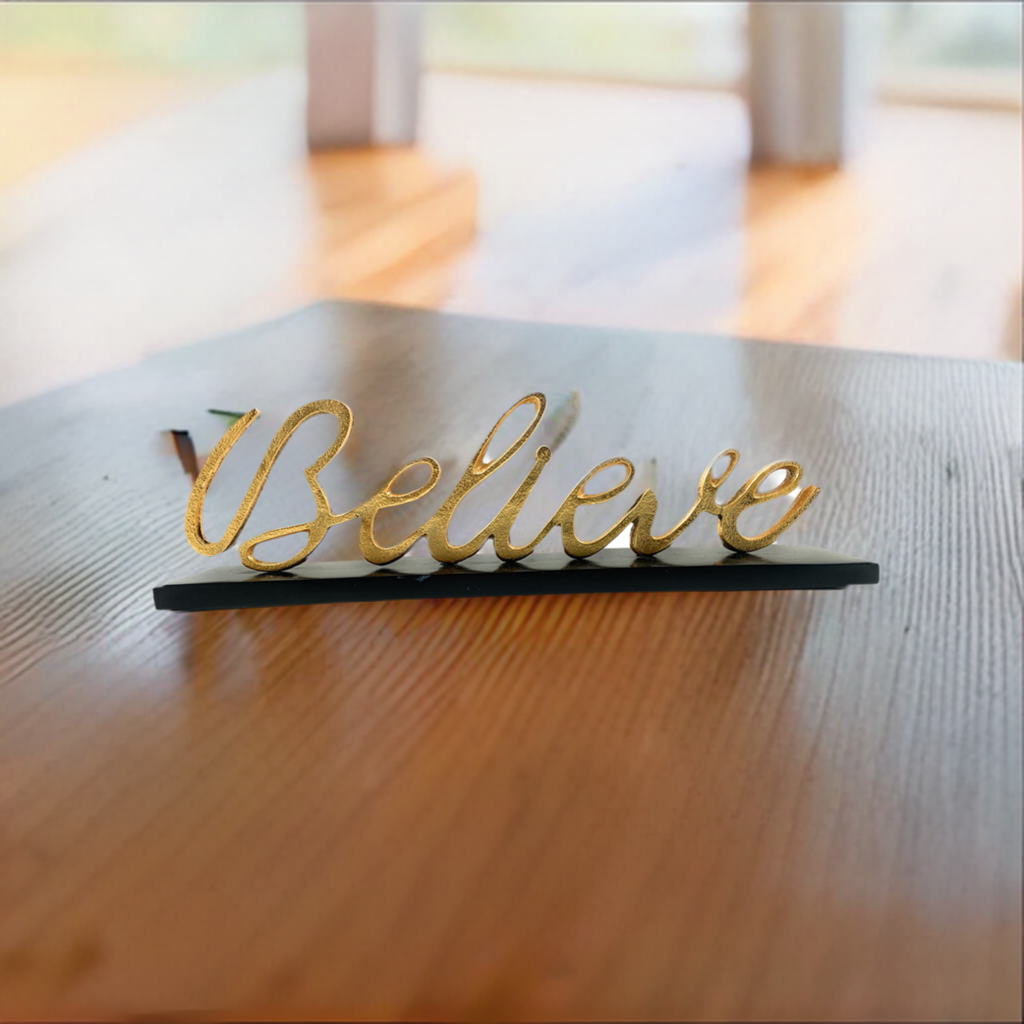 Gold script word 'Believe' on a black base, resting on a wood grain table with a blurred window background and soft natural lighting.