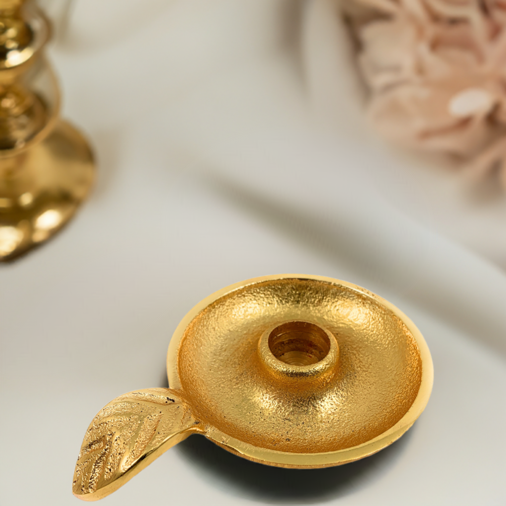 Gold-colored round candlestick holder dish with a textured surface and a decorative leaf-shaped handle on a light fabric background.