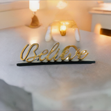 Gold-colored cursive word 'Believe' standing on a black base on a light-colored stone surface with warm glowing lights in the blurred background.