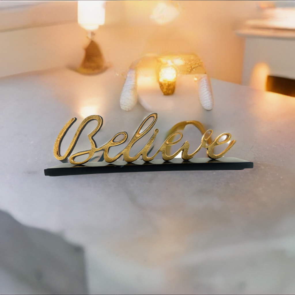 Gold-colored cursive word 'Believe' standing on a black base on a light-colored stone surface with warm glowing lights in the blurred background.